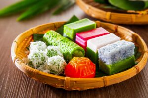 Assorted traditional Asian desserts arranged in a woven bamboo basket, featuring colorful layered kueh, pandan rolls, coconut-coated rice balls, and steamed sweets on banana leaves.