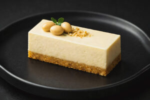 Rectangular soy cheesecake bar with a cookie crust, topped with whole soybeans and crushed nuts on a black plate.