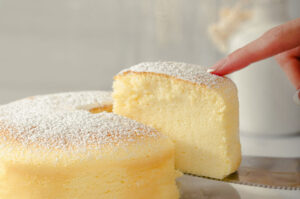 Soft and airy Japanese cheesecake dusted with powdered sugar as a hand gently presses the top, with a slice lifted on a cake server.