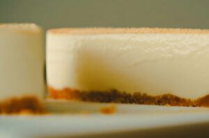 Close-up side view of a smooth no-bake cheesecake with a golden cookie crust on a white plate, showing its glossy creamy texture.
