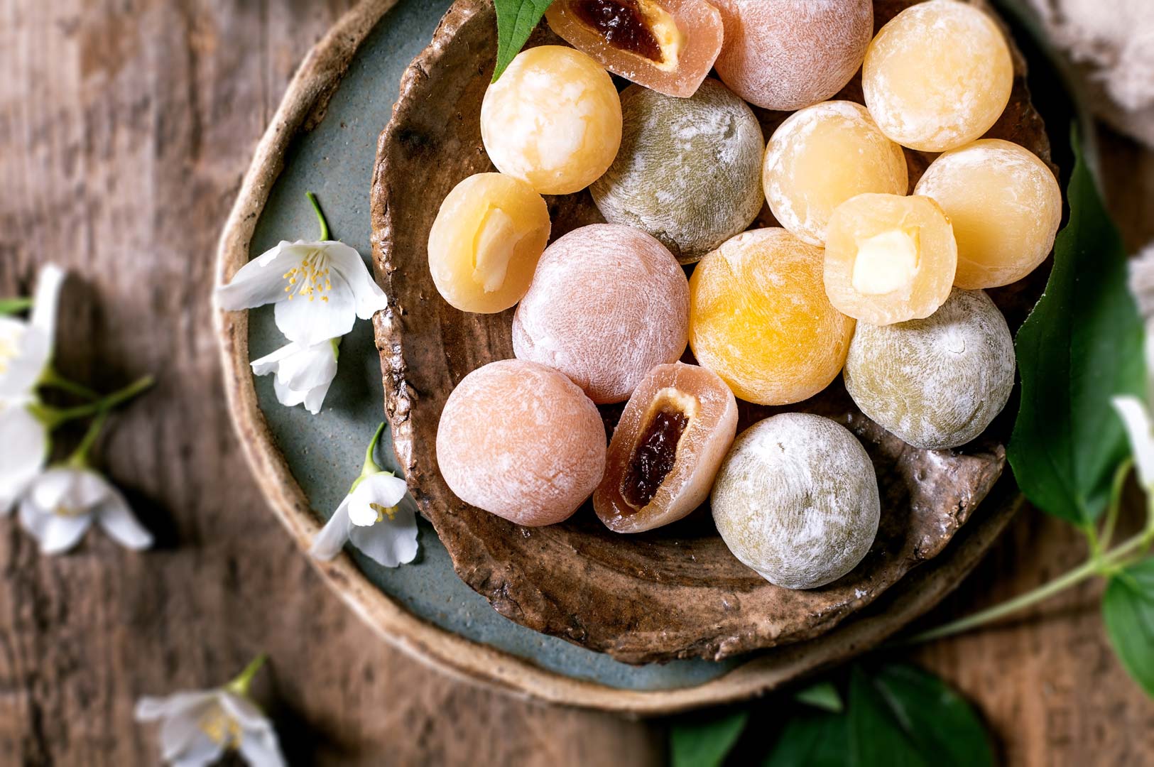 Assorted mochi in pastel colors displayed in rustic ceramic bowls, some filled with red bean and fruit paste, surrounded by green leaves and white blossoms.