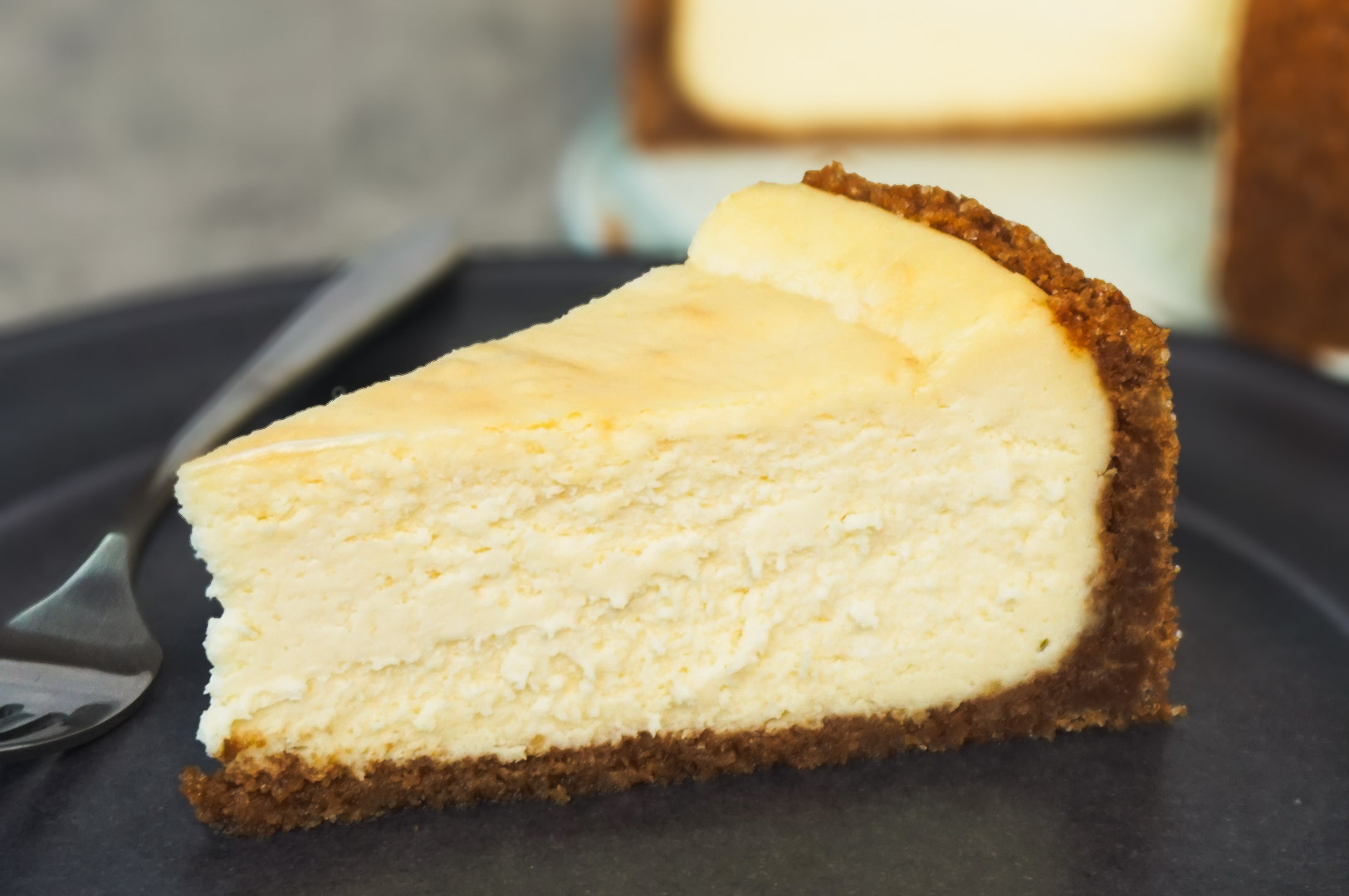 Classic baked cheesecake slice with a golden graham cracker crust on a dark plate.