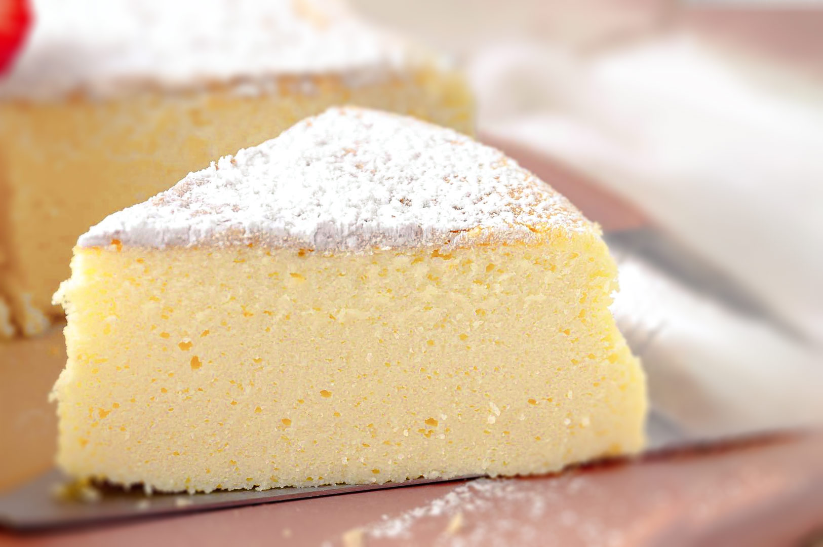 Slice of light and fluffy Japanese cheesecake dusted with powdered sugar, showing its airy texture and pale golden color on a soft-focus background.
