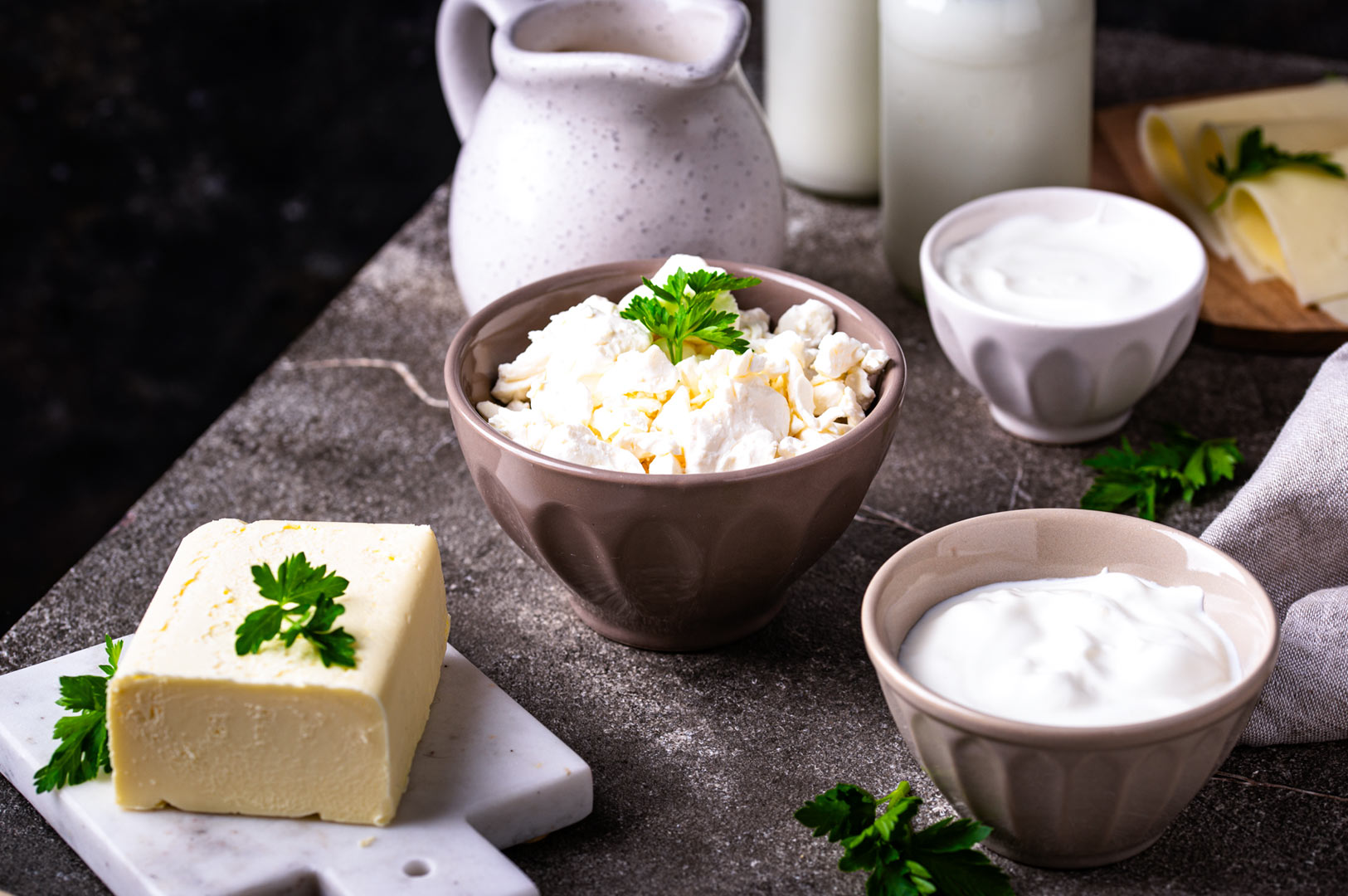 Assortment of dairy ingredients including cottage cheese, yogurt, butter, and milk displayed in ceramic bowls and pitchers on a rustic countertop.