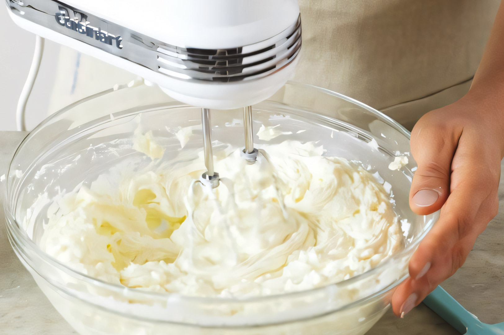 Electric hand mixer whipping a bowl of creamy cheesecake batter in a clear glass bowl held steady on a countertop