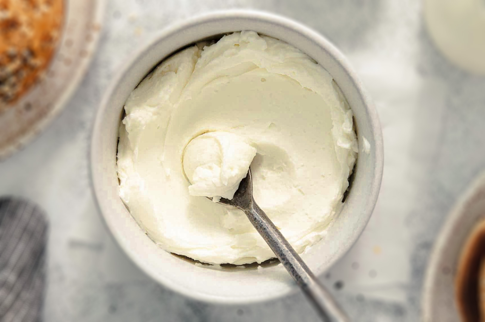 Creamy whipped cream cheese frosting in a ceramic bowl with a metal spoon, showing a smooth and fluffy texture.