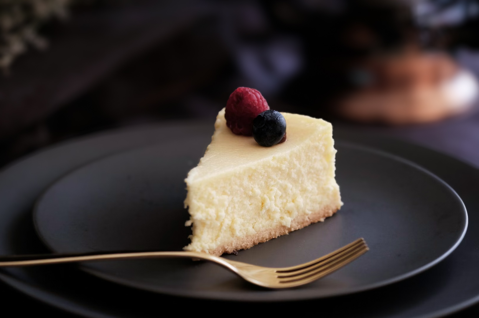 Slice of classic baked cheesecake on a dark plate topped with fresh raspberries and blueberries next to a gold fork.