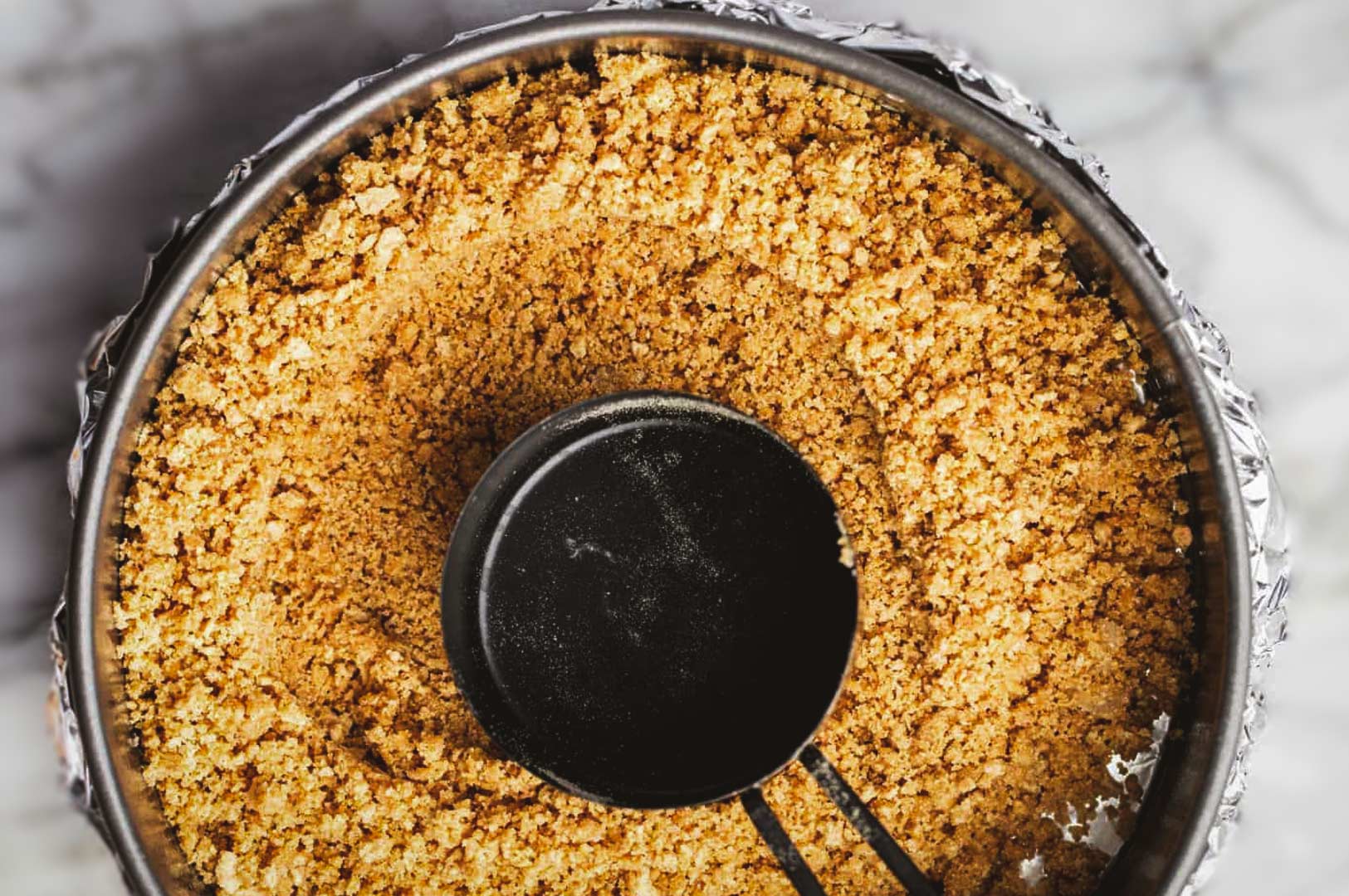 Crushed graham cracker crust being pressed evenly into the base of a springform pan using a metal measuring cup.
