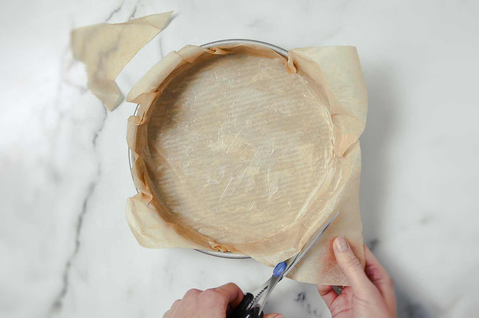 Hands trimming parchment paper around the edge of a round cake pan lined with baking paper on a marble surface.
