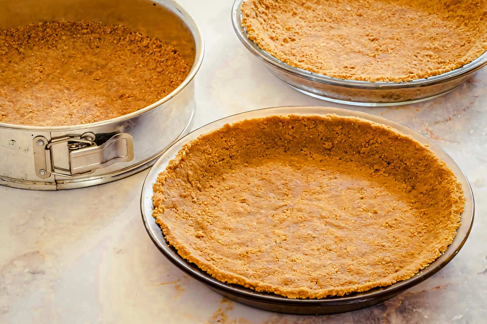 Three unbaked graham cracker crusts pressed into pie dishes and a springform pan, ready for filling on a light marble surface.