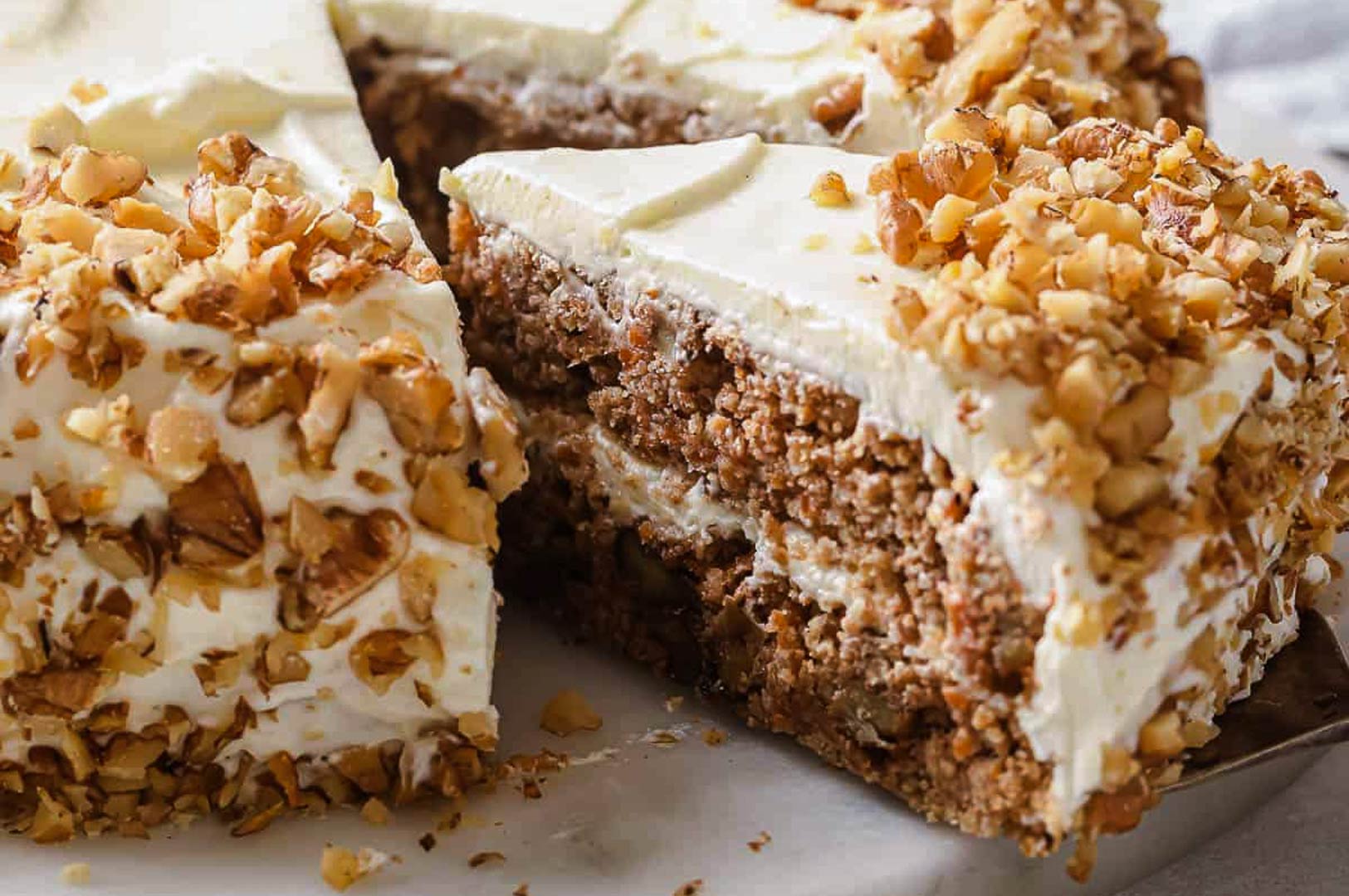 Moist carrot cake slice coated with chopped nuts and layered with creamy frosting, with a larger cake in the background.