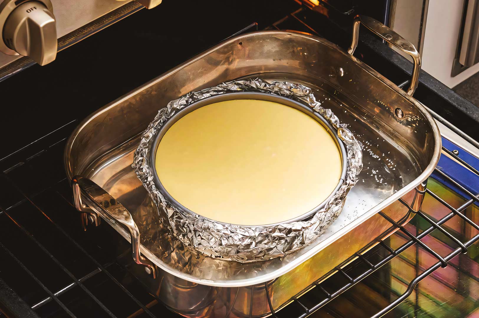 Unbaked cheesecake in a foil‑wrapped springform pan placed inside a large roasting pan filled with water for a bain‑marie, baking in the oven.