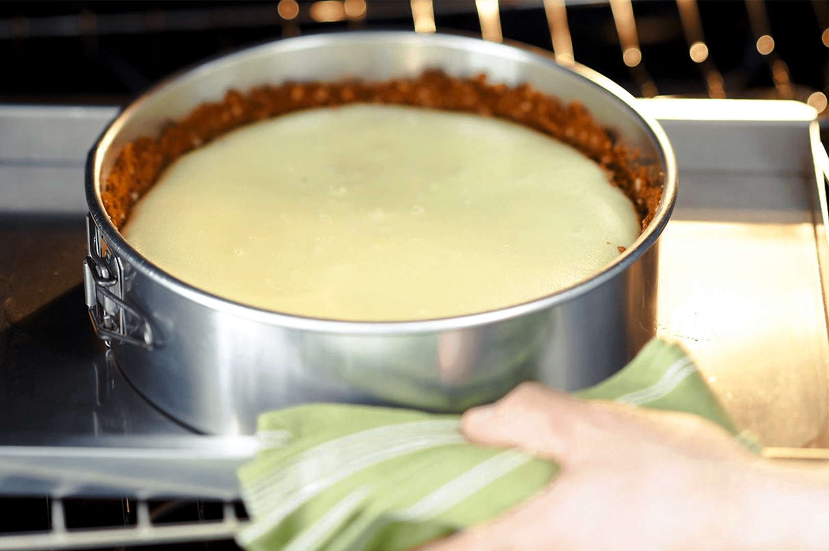 Cheesecake filling baking in a springform pan with a graham cracker crust as a hand uses a towel to adjust the oven tray.