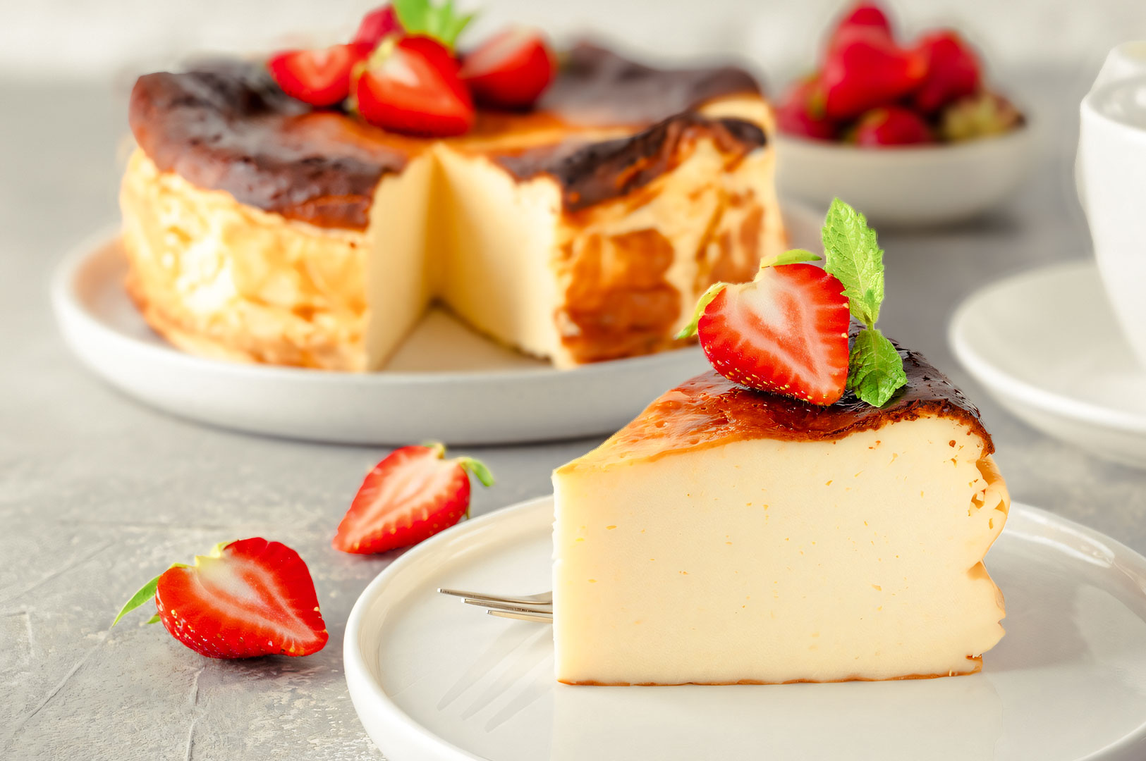 Slice of creamy Burnt Basque cheesecake topped with fresh strawberries, plated in front of the whole cheesecake on a light gray background.