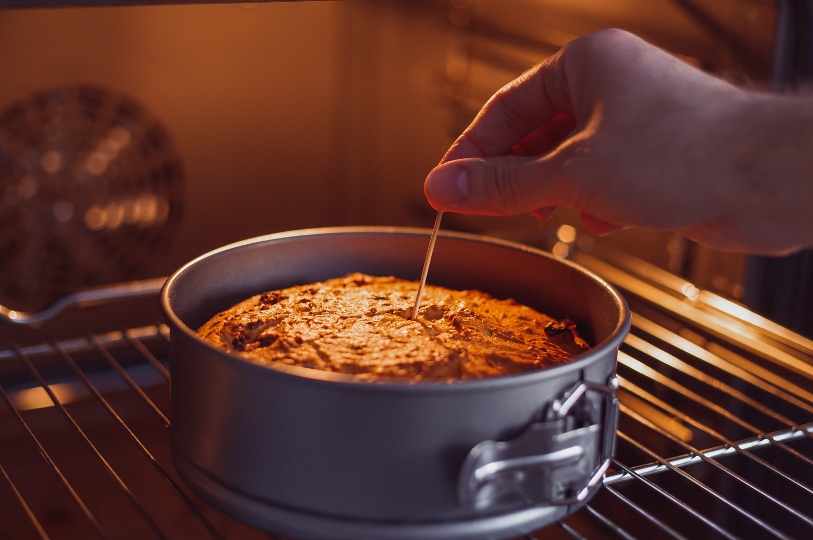 Hand inserting a toothpick into a freshly baked cake inside an oven to check for doneness, with the cake in a round springform pan on the oven rack.