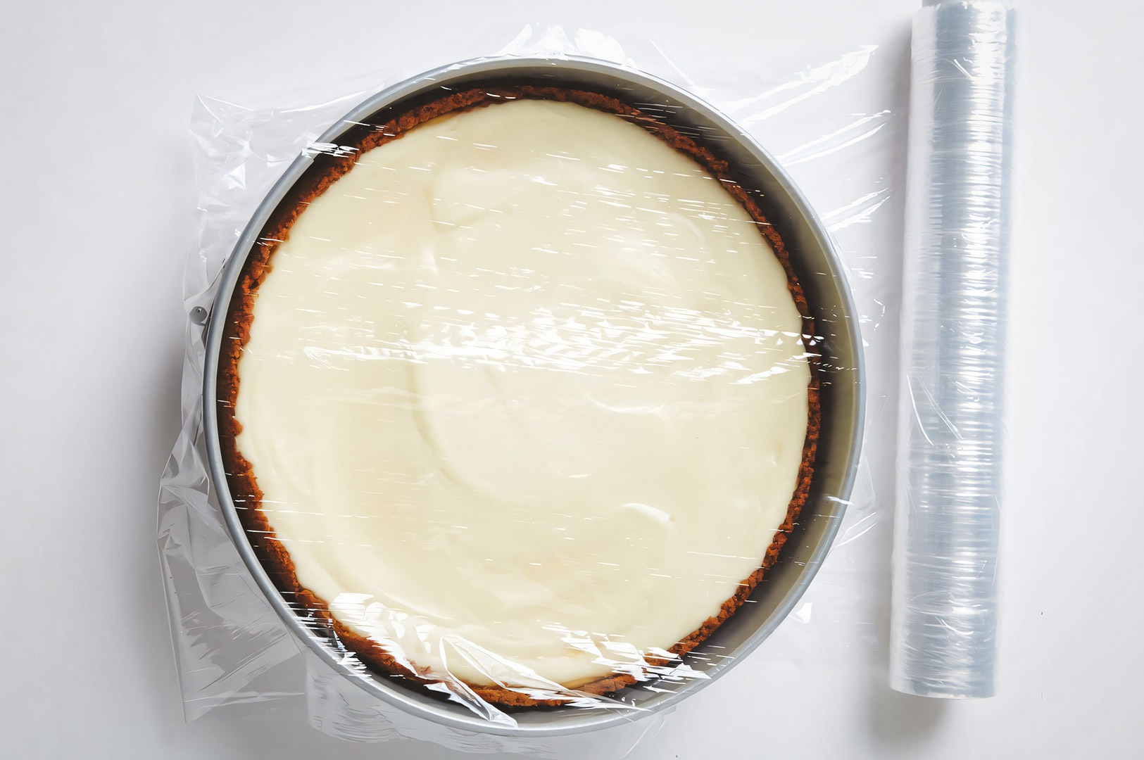 Chilled cheesecake in a springform pan covered tightly with clear plastic wrap, ready for storage or refrigeration on a white background.