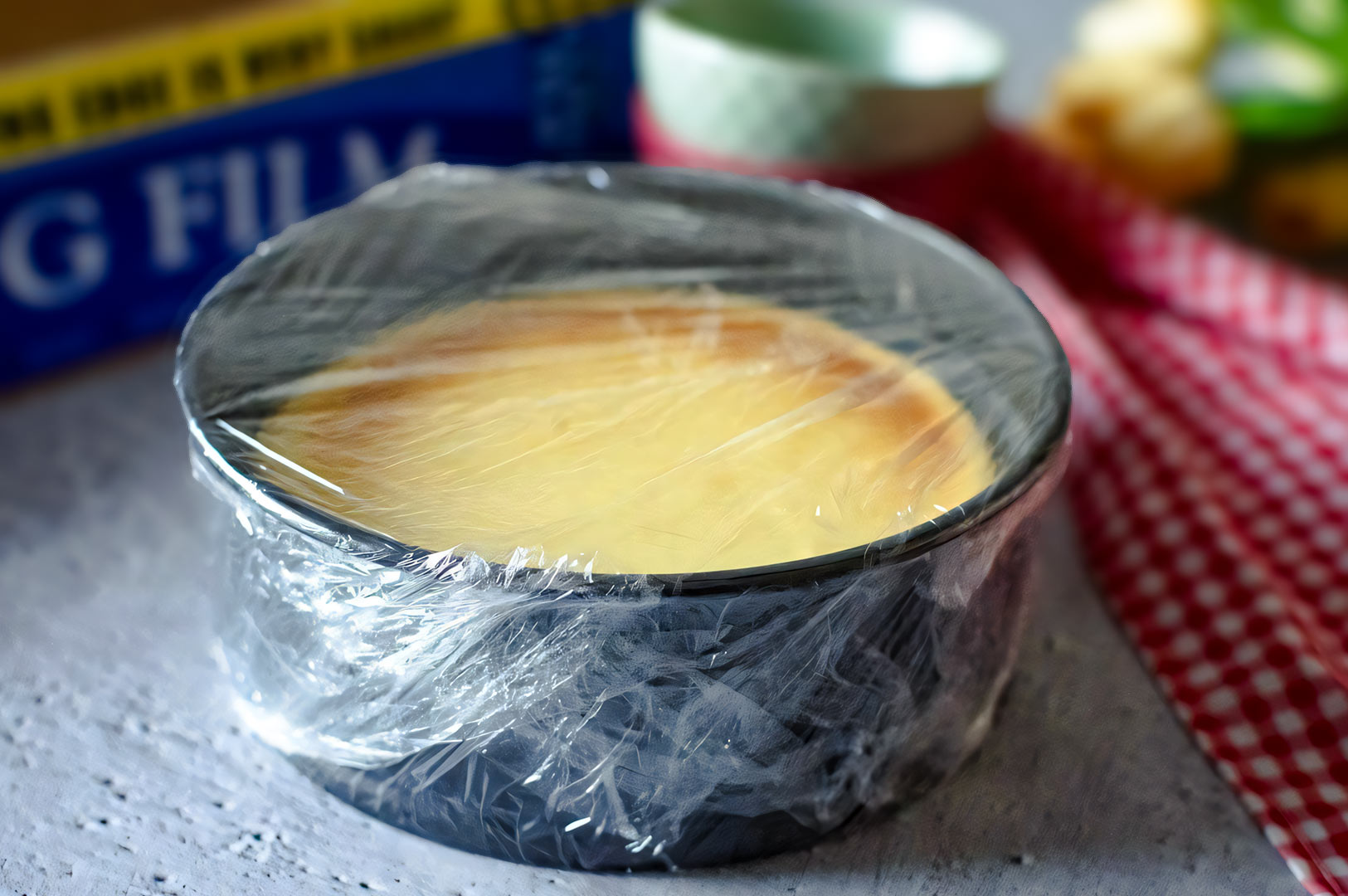 Freshly baked cheesecake in a round pan wrapped tightly in plastic cling film, cooling on a countertop with kitchen tools in the background.