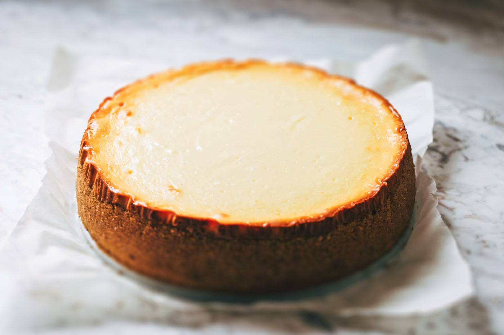 Freshly baked cheesecake with a lightly golden edge resting on parchment paper, cooling on a marble surface.