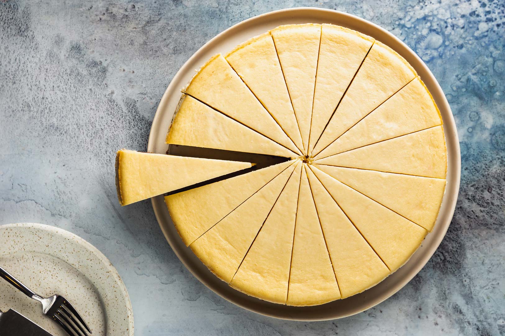 Whole baked cheesecake sliced into even portions with one slice pulled out, displayed on a round plate against a textured blue background.