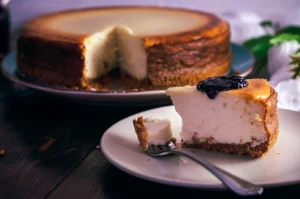 Slice of baked cheesecake on a plate topped with berry compote, with a whole cheesecake visible in the background.