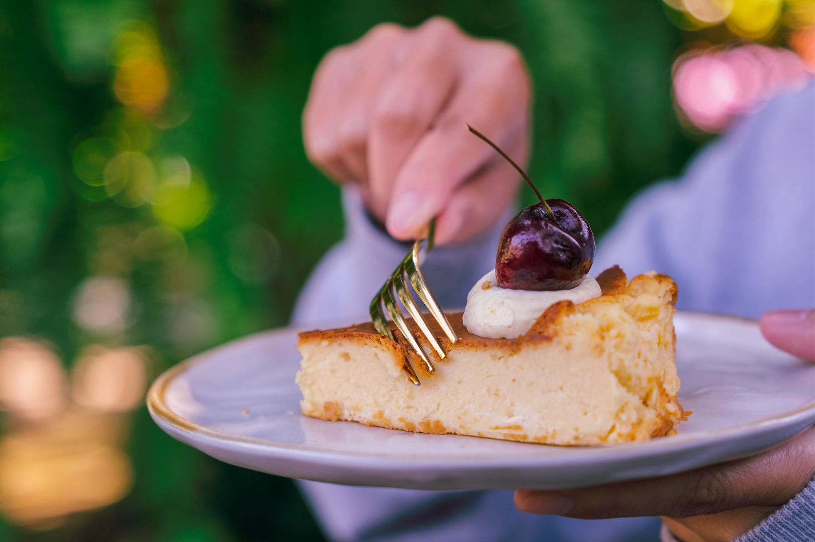 Slice of baked cheesecake served on a plate with whipped cream and a cherry, held outdoors in natural light