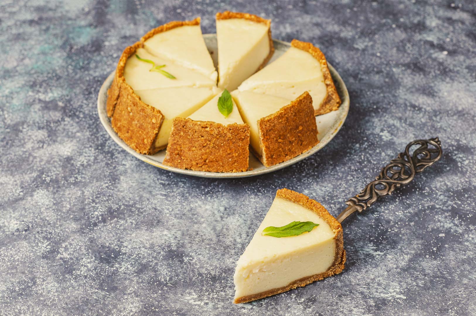 Whole creamy cheesecake with a graham cracker crust, sliced into even portions and garnished with fresh mint, displayed on a textured stone background.