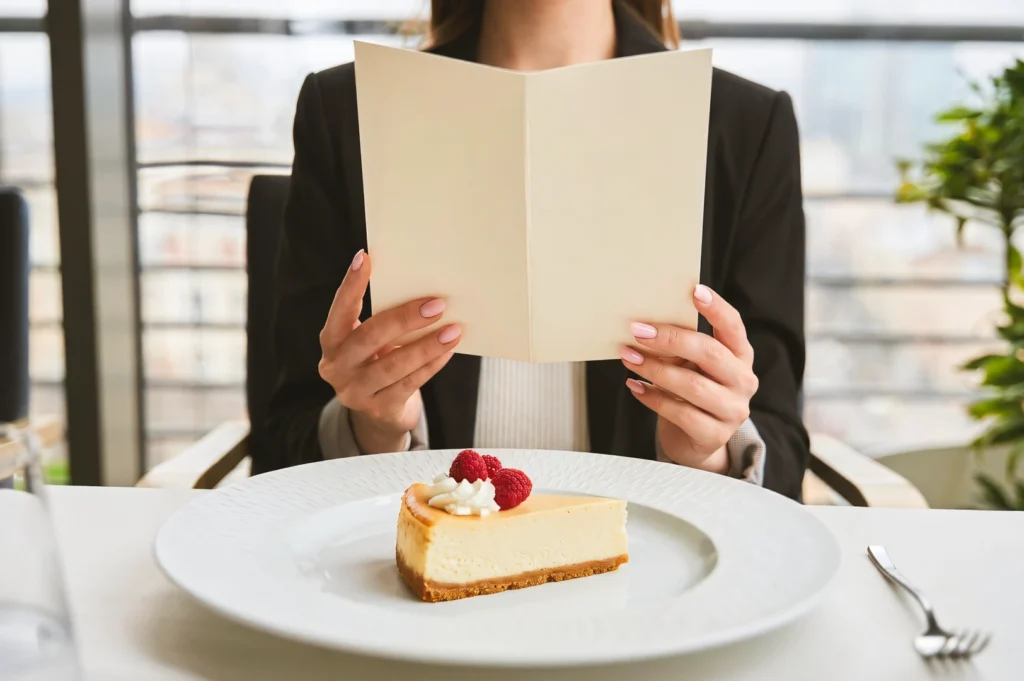 Eye‑level shot of a cheesecake slice garnished with raspberries on a white plate at a café table, with a menu held above the plate in a bright, modern dining setting.