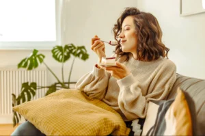 Eye‑level medium shot of a person seated on a sofa enjoying a dessert from a small plate, styled in a cozy indoor living space with natural light and houseplants.