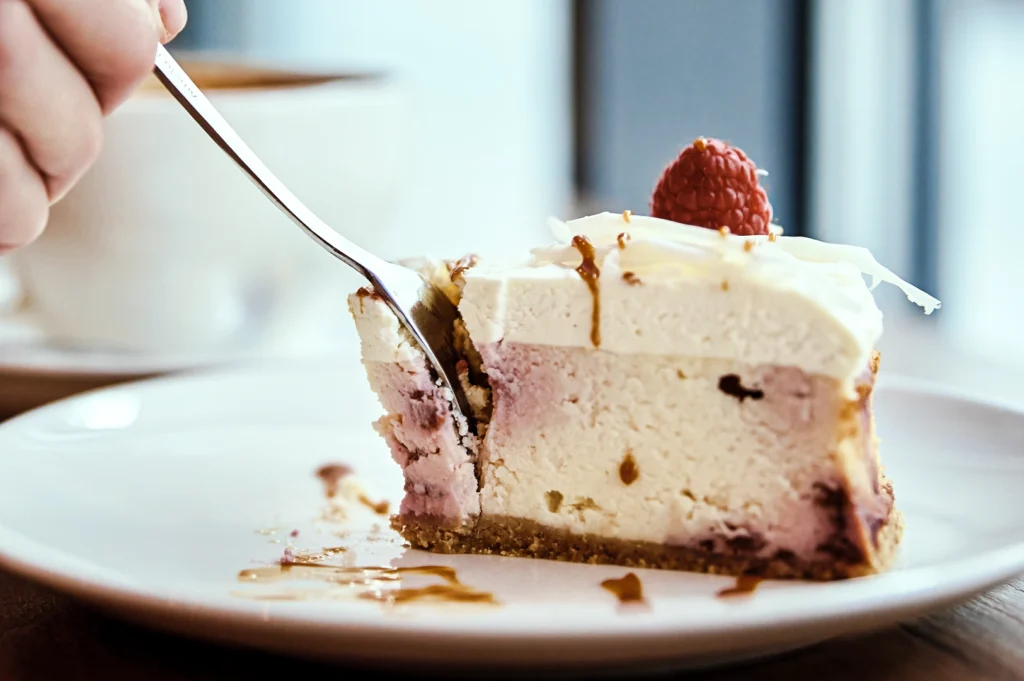 Slice of raspberry‑topped cheesecake with a graham crust being cut with a fork, featuring creamy layers and caramel drizzle on a white plate.