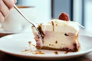 Slice of raspberry‑topped cheesecake with a graham crust being cut with a fork, featuring creamy layers and caramel drizzle on a white plate.