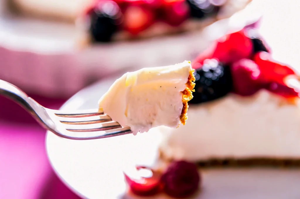 Close‑up of a fork holding a creamy cheesecake bite with graham cracker crust, blurred cheesecake slice in background.