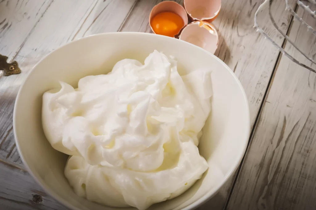 Top-down shot of a bowl filled with glossy whipped egg whites on a rustic wooden surface, with cracked eggshells and yolks nearby, illustrating baking preparation.
