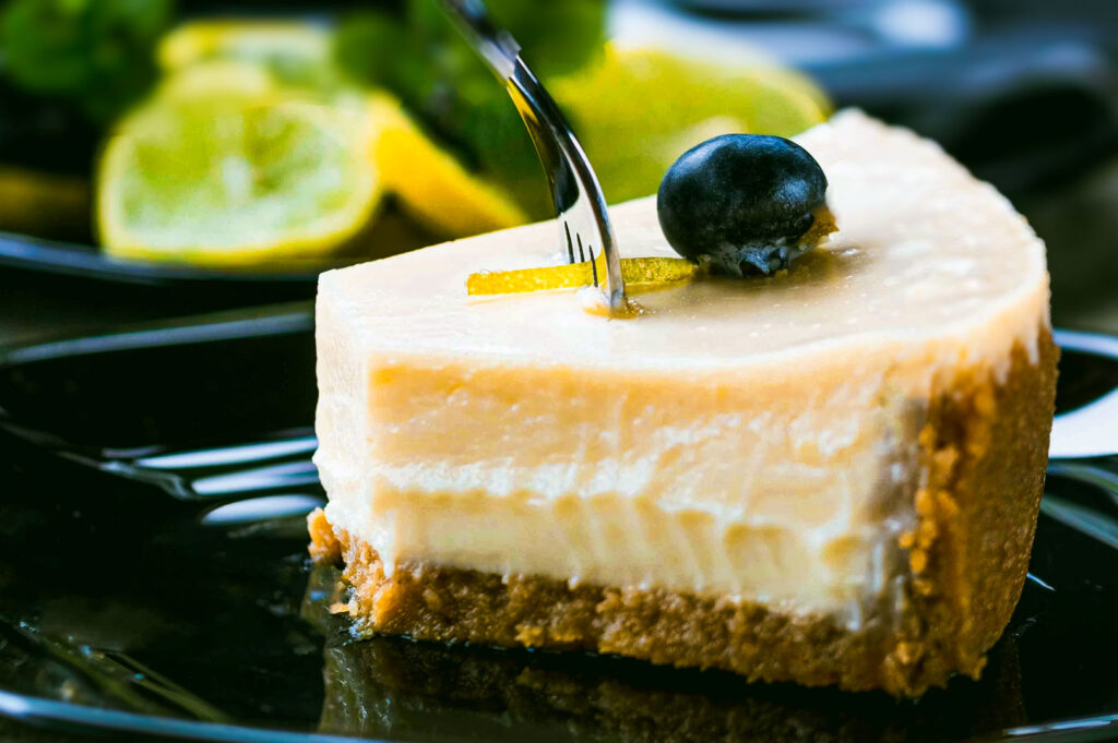 A slice of New York style cheesecake with a graham crust, topped with a blueberry and lemon zest, served on a glossy black plate.