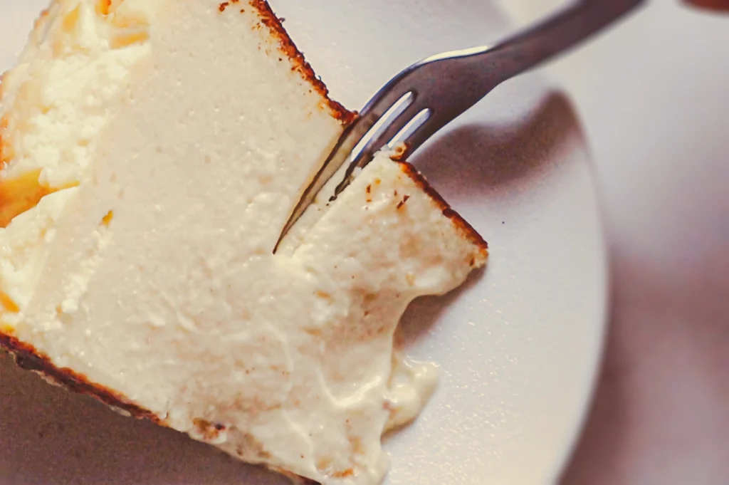 Close‑up of a creamy cheesecake slice on a white plate with a fork, highlighting the smooth texture and rich baked cheesecake filling