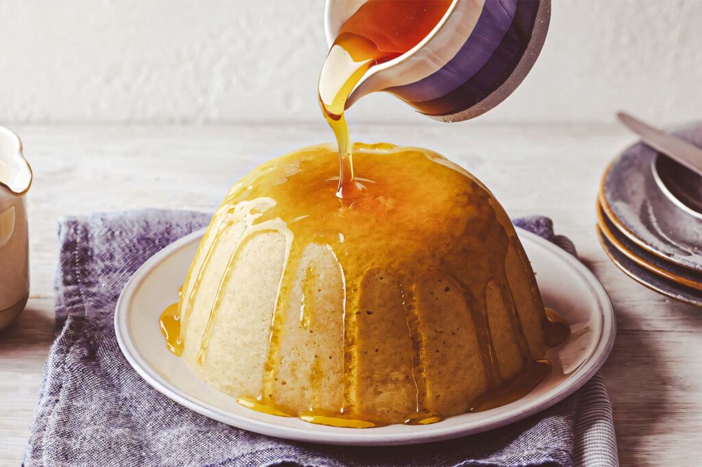 Golden syrup being poured over a warm steamed sponge pudding on a white plate, creating a glossy caramel glaze