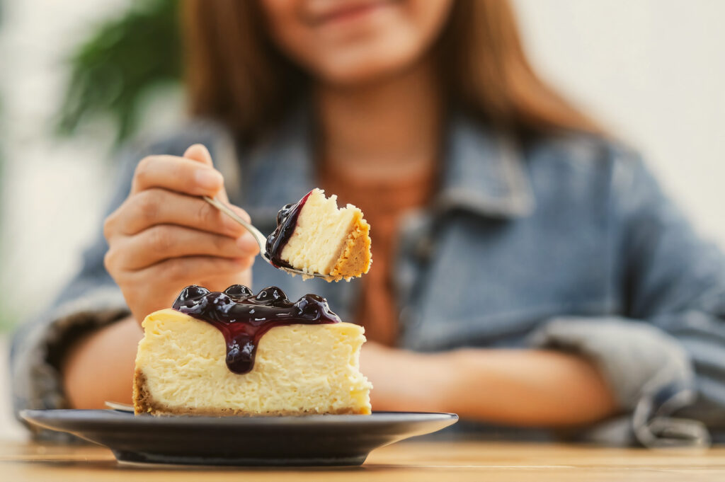 Close‑up of a blueberry cheesecake slice topped with glossy blueberry compote, placed on a dark plate with a fork lifting a bite