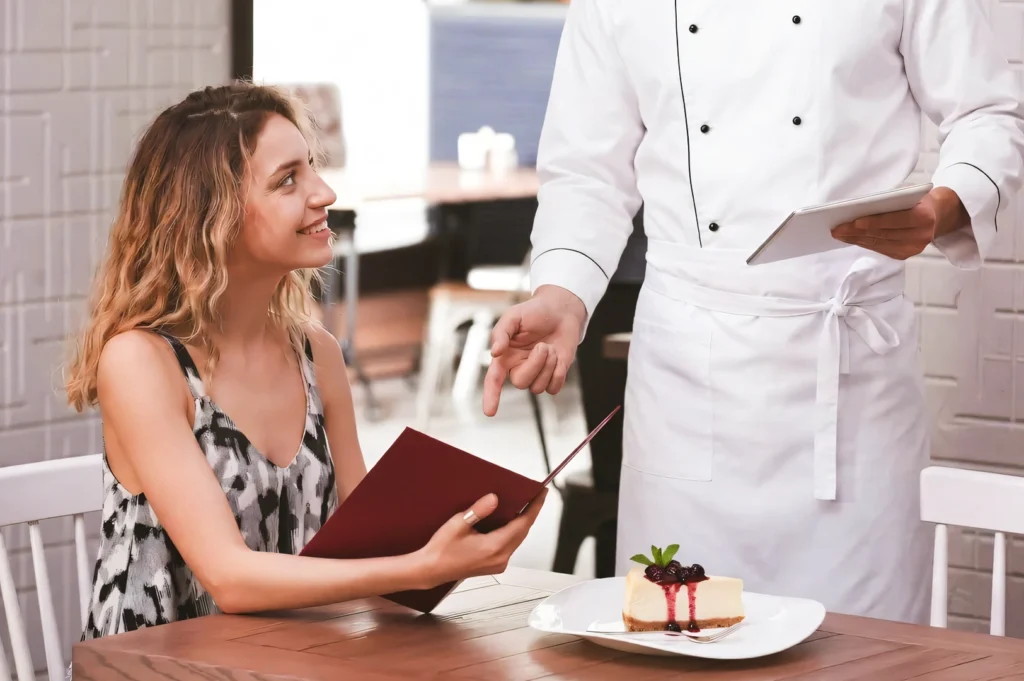 Mid‑angle shot of a restaurant table where a server presents a plated cheesecake dessert while a diner reviews a menu, highlighting a refined dining experience and table‑side service.