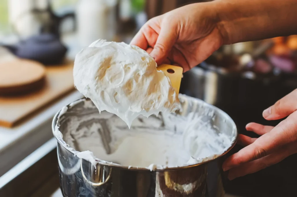 Close-up kitchen shot of thick whipped cheesecake batter lifted from a metal mixing bowl with a spatula, showing smooth texture and baking preparation details.