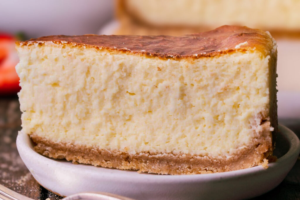 Thick slice of classic New York cheesecake with a golden top and graham cracker crust served on a small plate.