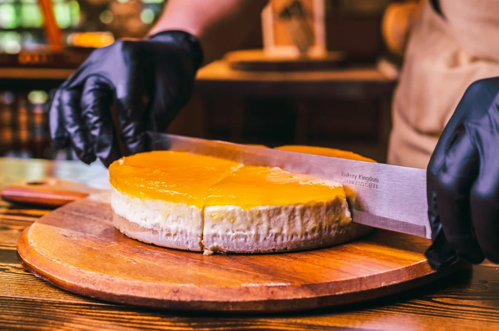 Chef slicing a creamy mango cheesecake with a glossy topping on a wooden board using a sharp kitchen knife