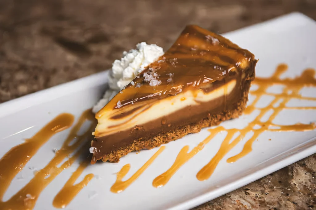 Three-quarter angle shot of a sliced salted caramel cheesecake with a biscuit crust, topped with glossy caramel sauce and whipped cream, plated on a rectangular dish for dessert presentation.