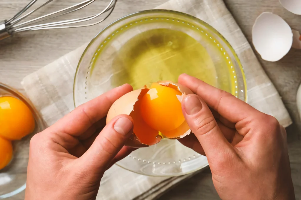 Top-down shot of hands cracking a fresh egg over a clear glass mixing bowl, with visible egg yolk and whites, surrounded by baking tools on a light kitchen surface.