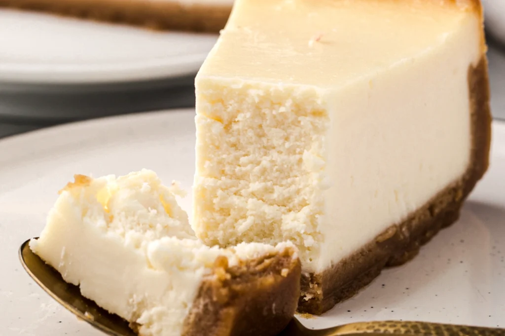 Classic New York‑style cheesecake slice with a thick graham cracker crust and a rich, dense filling, served with a spoonful cut out.