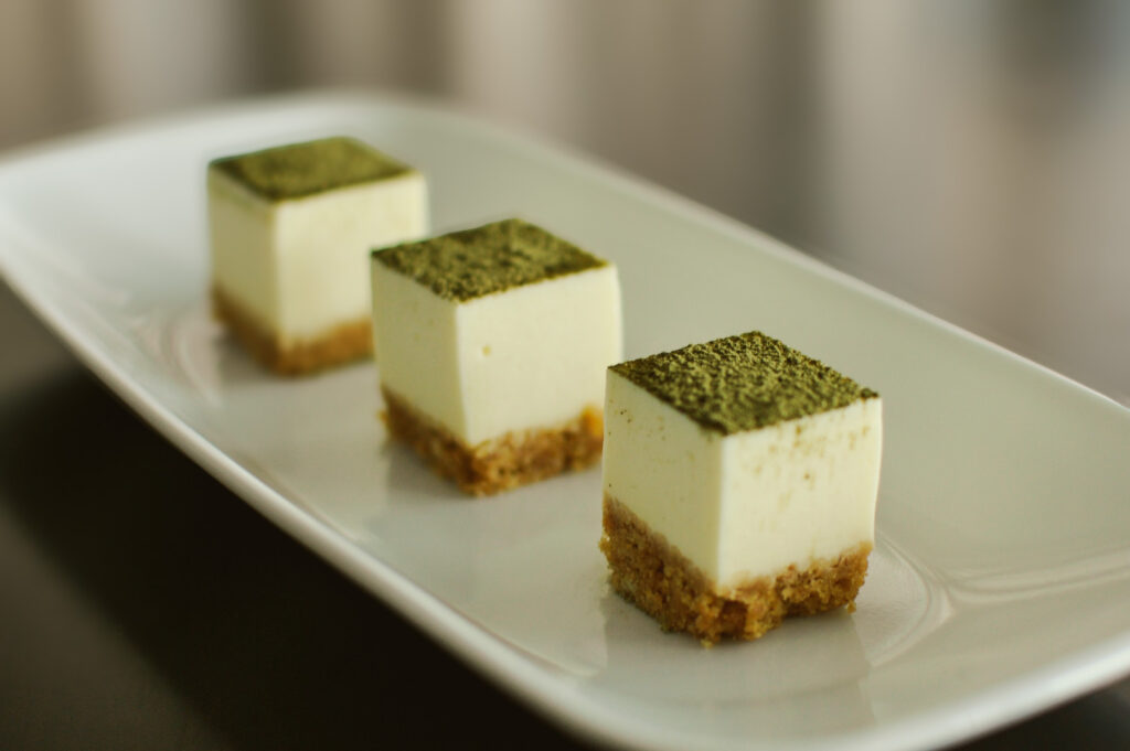 Three square matcha cheesecake bites arranged on a white rectangular plate, featuring a layered graham crust and matcha‑dusting topping