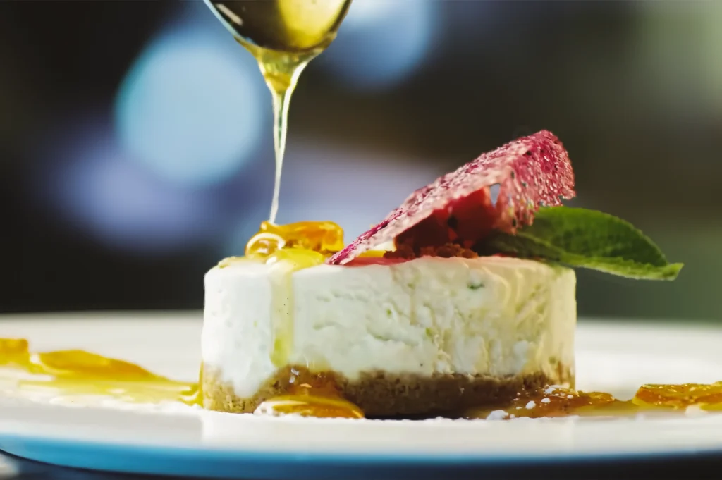 Close-up shot of a creamy cheesecake slice topped with fruit and herbs as golden honey is drizzled from above, emphasizing texture, freshness, and gourmet dessert presentation.