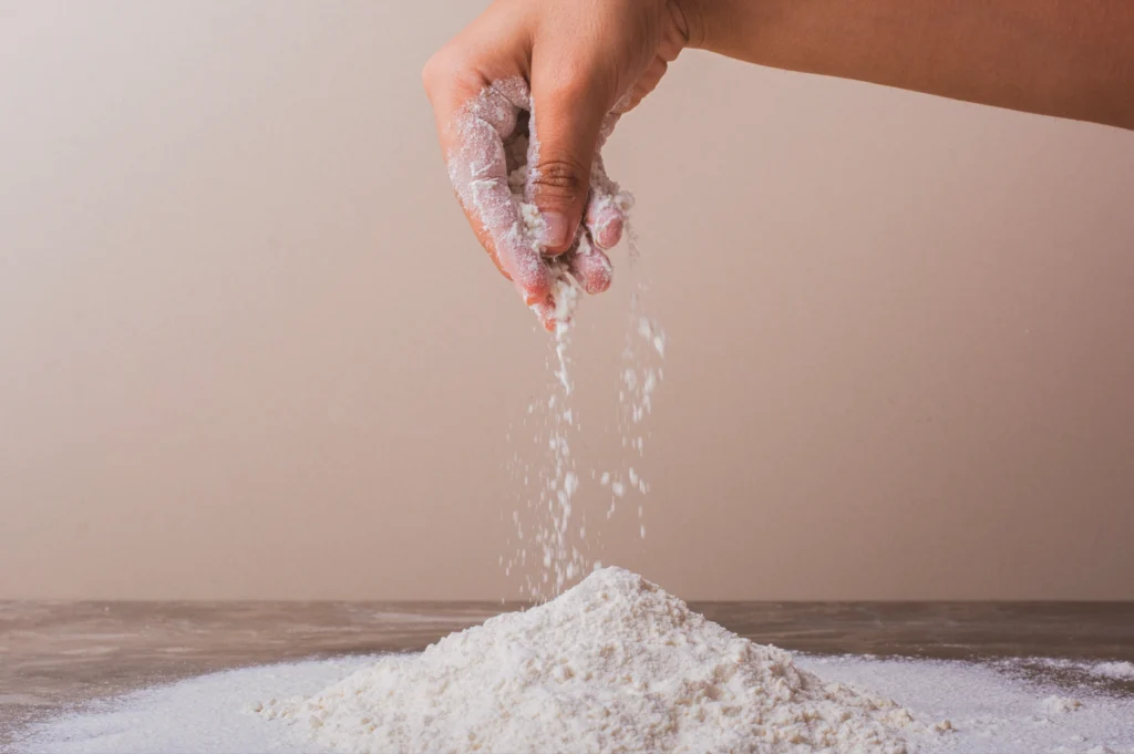 Mid‑angle shot of a hand sprinkling flour over a small mound on a wooden surface, capturing falling flour particles against a soft neutral background.