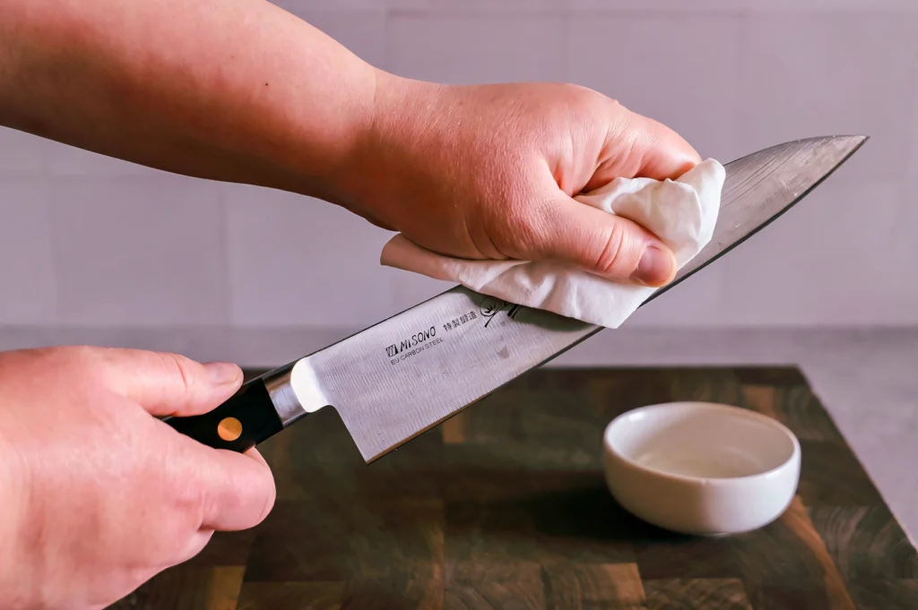 Hands carefully wiping a chef’s knife with a cloth on a wooden cutting board