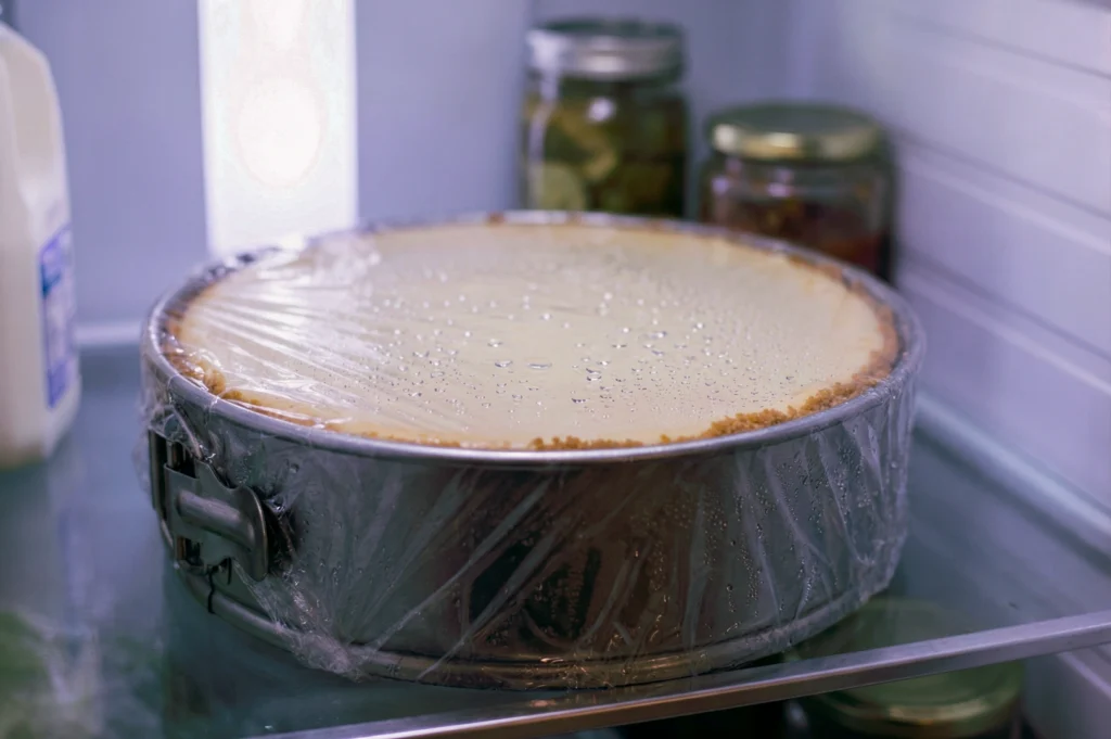 New York style cheesecake wrapped in plastic inside a refrigerator, resting in a springform pan while cooling.