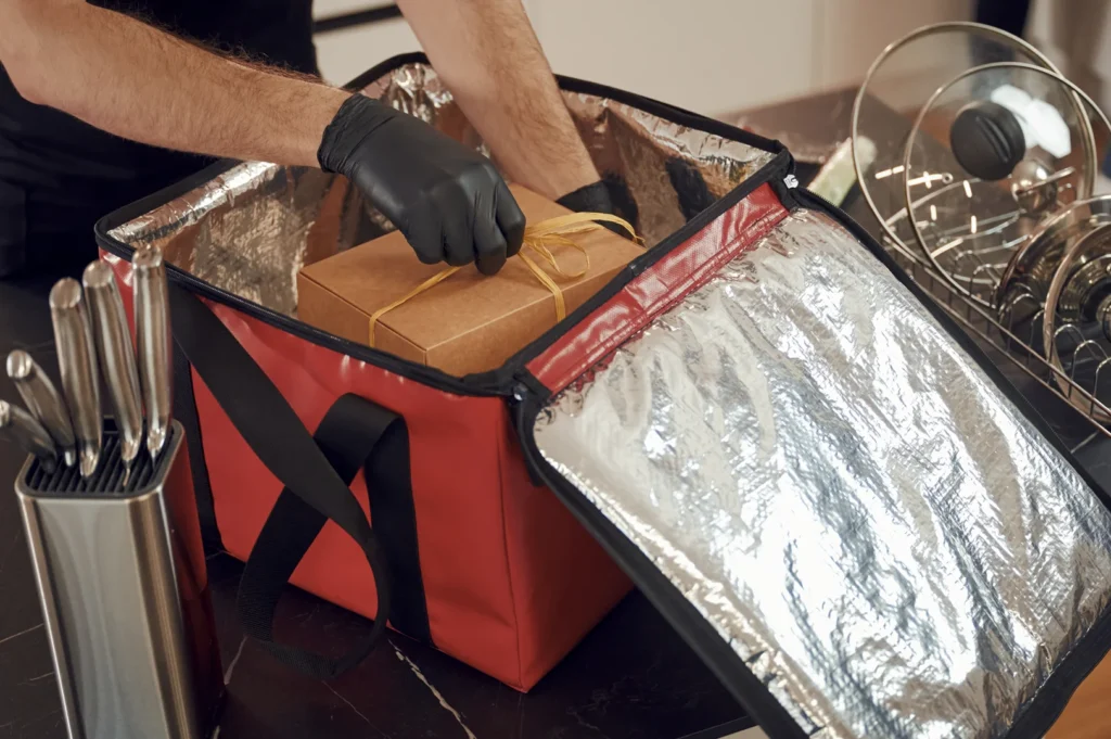 Insulated food delivery bag being packed with a tied cardboard box, handled by someone wearing black gloves