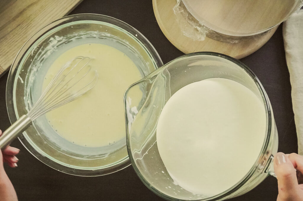 Top‑down view of two glass bowls containing cheesecake batter, with a whisk and a jug of milk being prepared for baking